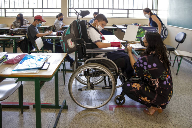 Em uma sala de aula, uma mulher de roupa escura está agachada em frente a um garoto em uma cadeira de rodas