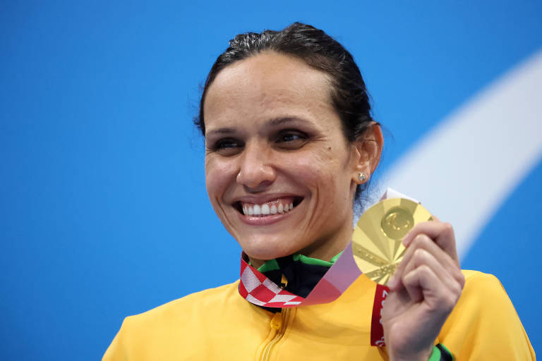 Nadadora Carol Santiago segura sua medalha de ouro das Paralimpíadas ao lado de seu rosto, e sorri