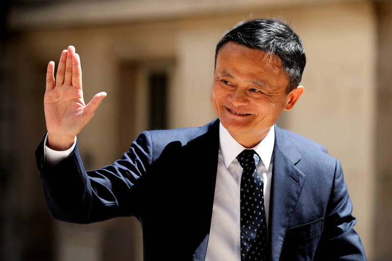 Jack Ma usa um terno preto, camisa branca e cabelo curto preto; ele levantar a mão direita saudando em direção da câmera