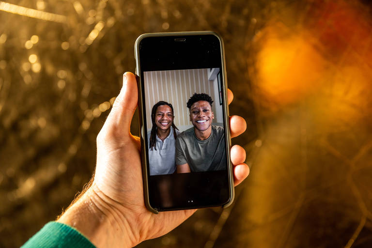 Uma mão segura um celular e na tela aparece dois jovens negros sorrindo