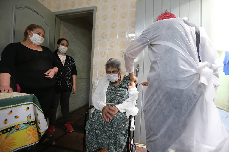 Na sala de uma casa, um enfermeiro aplica vacina no braço de uma idosa que está sentada em uma cadeira. De pé, duas mulheres observam a cena.