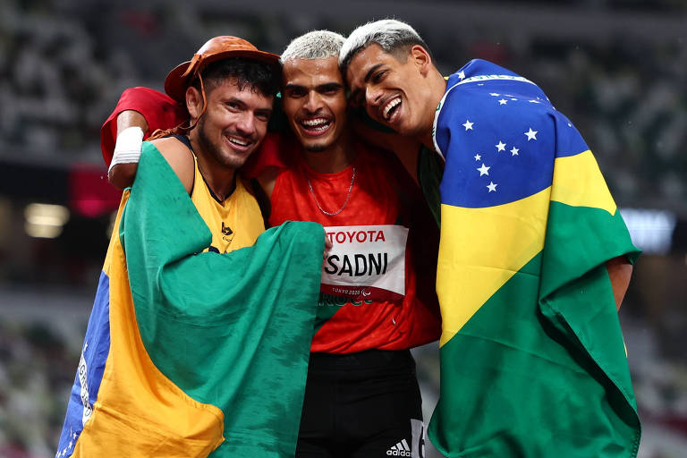 Os três atletas abraçados e sorrindo, com os brasileiros enrolados na bandeira do país
