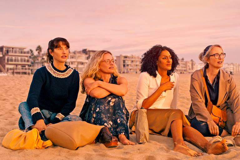 Quatro atrizes sentadas na areia de uma praia, com roupas casuais, olhando para ao horizonte