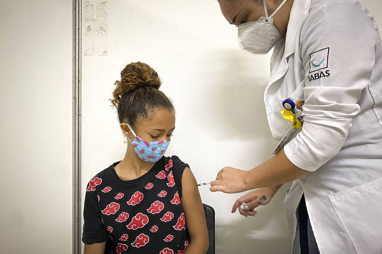 Menina de cabelo preso, usando máscara, está sentada e olha para seu braço enquanto enfermeira aplica vacina