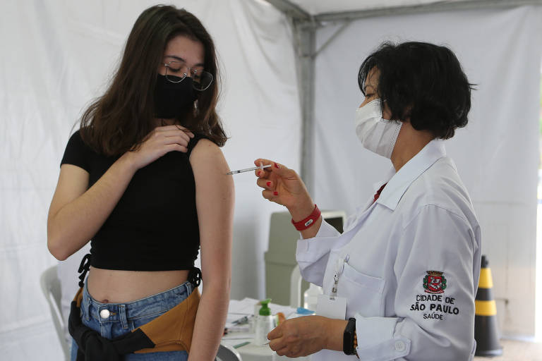 Jovem de cabelo longo, blusa preta, calça jeans recebe uma injeção no braço aplicada por uma enfermeira de jaleco branco. Ambas usam máscaras