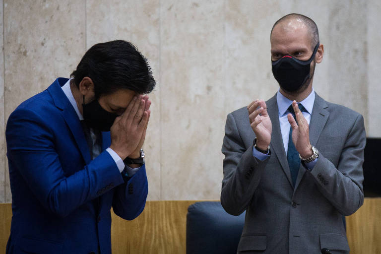 Ricardo Nunes, à esquerda, junta as mãos à frente do rosto e inclina a cabeça, como se estivesse rezando. Ao seu lado, Bruno Covas aplaude.