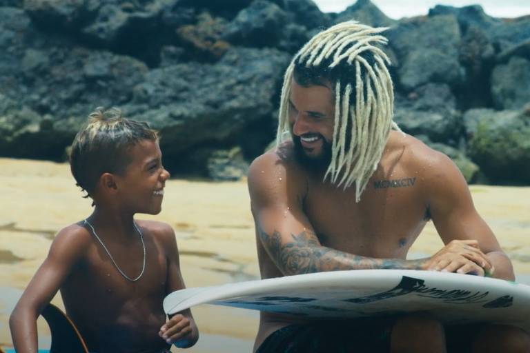 Cena do documentário "A Curiosa História de Italo Ferreira" mostra o surfista Italo Ferreira ao lado de uma criança, os dois sentados, com as pranchas de surfe no colo, em uma praia de Baía Formosa