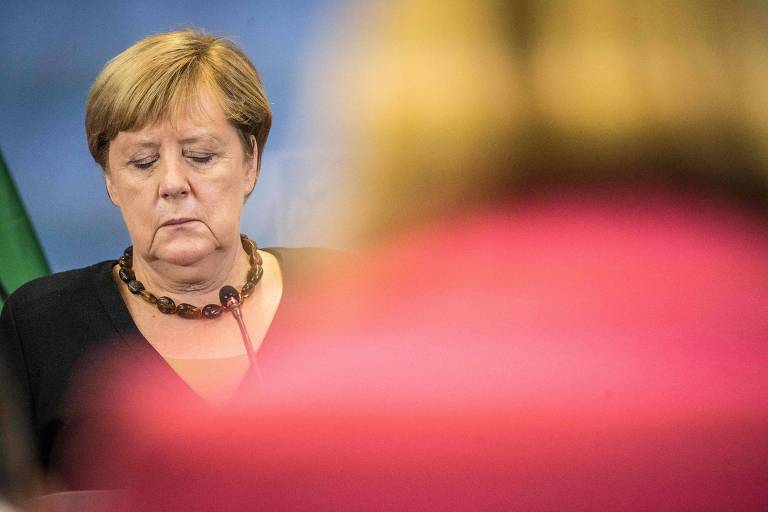 De blusa preta com gola em V e um colar de bolas pretas, Merkel olha para baixo