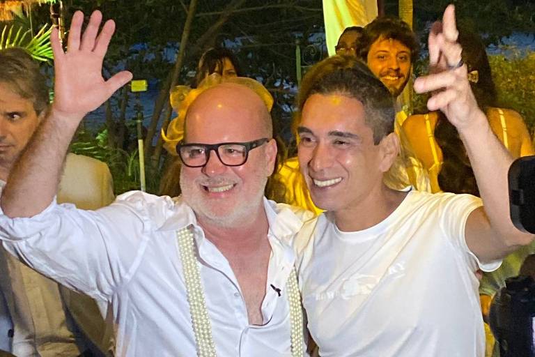 Dois homens brancos vestindo roupas brancas abraçados em meio a uma festa