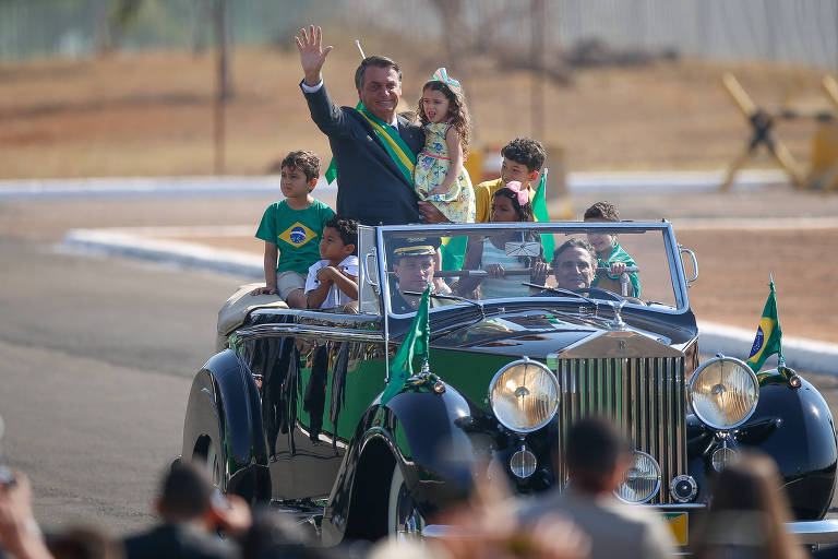 Com uma criança amparada no seu braço esquerdo, o presidente Jair Bolsonaro chega em carro aberto, dirigido pelo piloto ex-piloto de Fórmula 1 Nelson Piquet, para a cerimônia de hasteamento da bandeira no Palácio da Alvorada