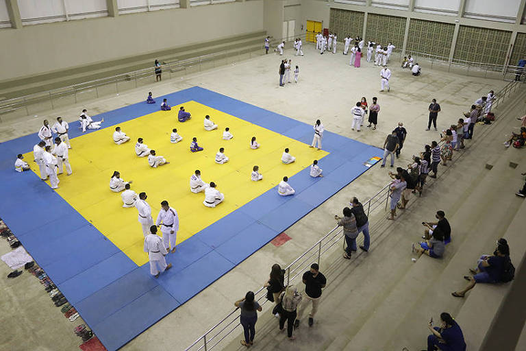 Vista geral do interior do ginásio Sarah Menezes, em Teresina, com pessoas sentadas no tatame antes de aula de judô