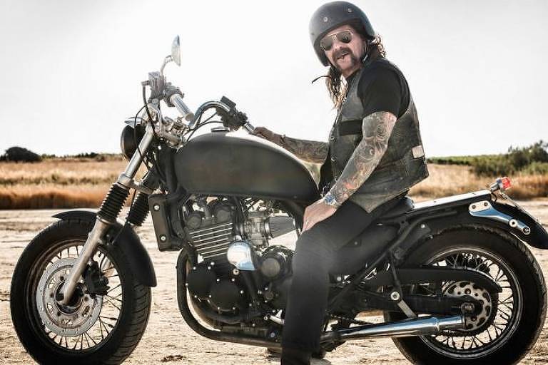 Foto mostra homem vestindo preto, de capacete e com braço tatuado montado numa moto num local de vegetação seca