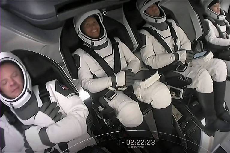 Quatro pessoas sentadas com roupas de astronauta brancas dentro de uma nave com paredes e assentos cor de cinza