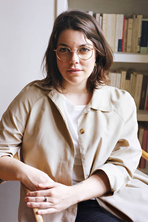 mulher branca de cabelos lisos castanhos e óculos sentada diante de estante