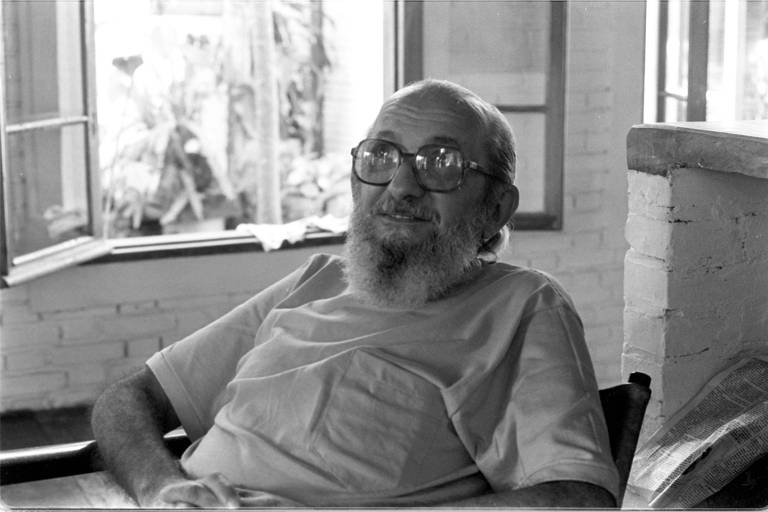 Pàulo Freire, de barba e óculos, sentado, com as mãos sobre a barriga, olha para o fotógrafo; ao fundo, uma janela aberta