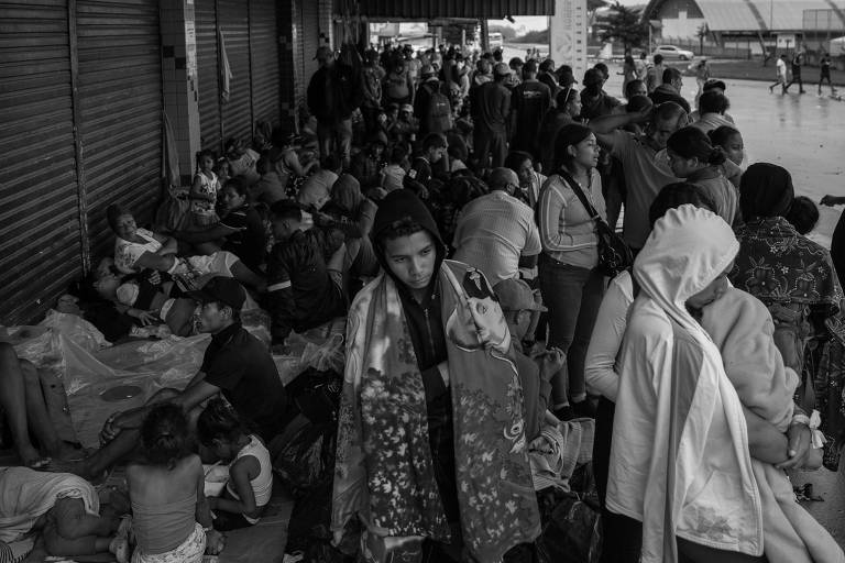 Dezenas de pessoas reunidas sob a marquise de uma galeria de lojas, numa rua de Pacaraima (RR). As pessoas estão com cobertores, algumas em pé, a maioria deitadas no chão. Há homens, mulheres, jovens e crianças.