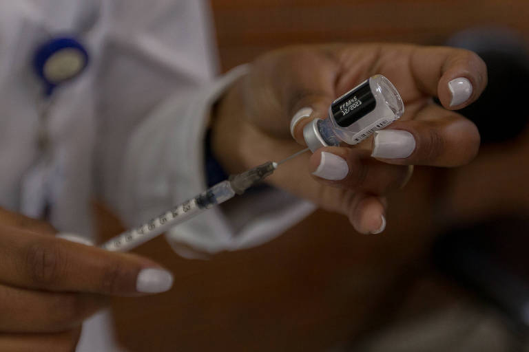Imagem destaca mãos manipulando uma seringa e um vidro de vacina