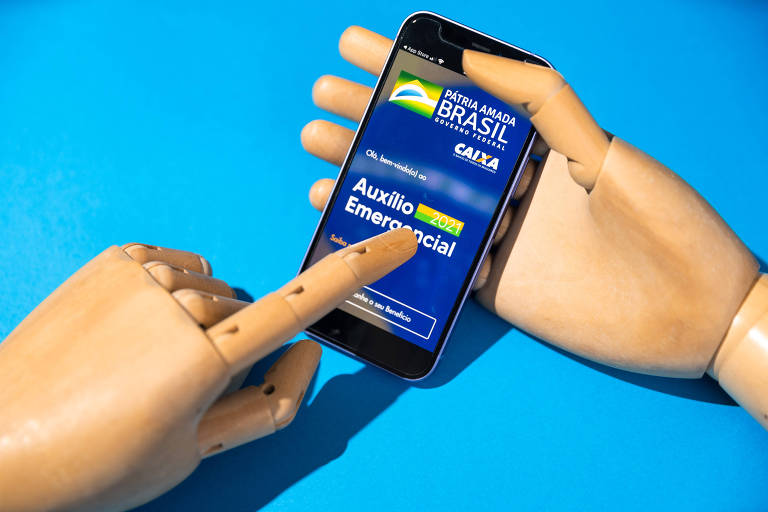 Fotografia colorida de duas mãos articuladas de madeira segurando um celular onde está aberta uma tela azul onde se lê "Auxílio emergencial" e no topo, "Pátria amada Brasil".