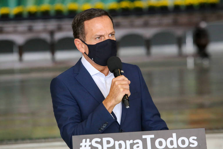Foto mostra o governador João Doria de máscara, falando em um microfone em um tablado escrito SP para todos.