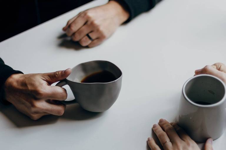 Duas mãos seguram uma xícara de café cada, apoiadas em uma mesa