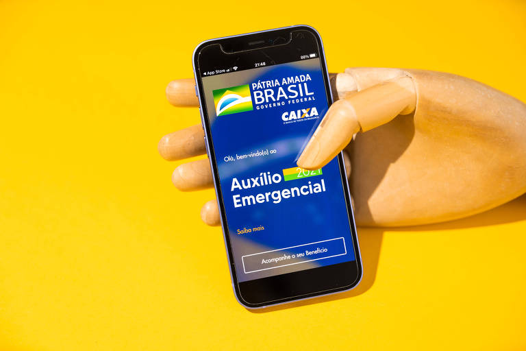 Foto mostra um fundo amarelo com uma mão articulada de madeira segurando um celular, onde uma tela azul do aplicativo do auxílio emergencial está aberta.