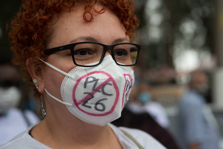 Foto mostra o rosto de uma mulher de pele clara, cabelo curto encaracolado ruivo, óculos quadrados e pretos, usando uma mascara branca, que cobre o nariz e a boca, escrito PLC 26 com um símbolo de proibido em cima.