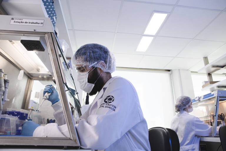 Homem usando máscara, jaleco, touca e luvas de proteção utiliza instrumento em um laboratório. Ao fundo, outra cientista aparece sentada.
