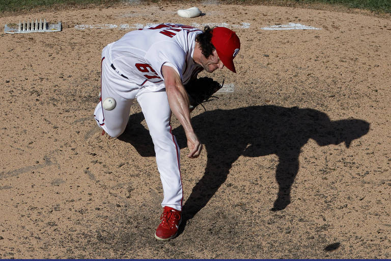 Jogador de beisebol arremessa bola, com a perna esquerda bem a frente do corpo e o braço direito também; ao lado dele, a sua sombra está nítida