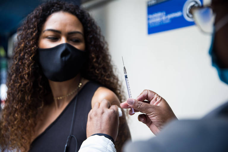 Mulher negra, de máscara, observa profissional de saúde segurar seringa com a vacina