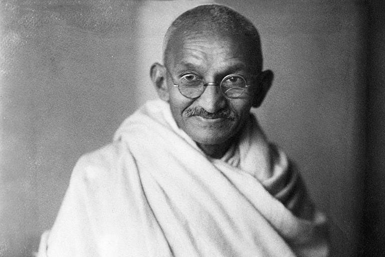 Mahatma Gandhi aos 62 anos, careca, com óculos redondos, bigode e uma túnica branca, esboça um sorriso para a foto