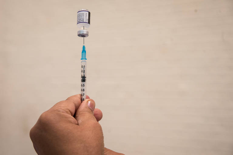 Frasco de vacina da Pfizer apoiada sobre seringa, segurada por profissional de saúde