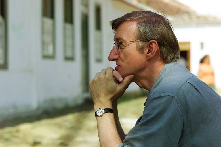 Perfil de um homem branco de cabelos grisalhos, óculos e camisa azul clara, com a mão no queixo usando um relógio preto em uma rua de pedras com casas brancas antigas de janelas grandes