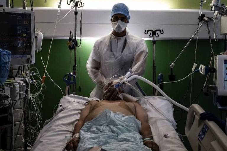 Médico em pé, paramentado com roupas de proteção e máscara, cercado por maquinário médico e em frente à maca de um paciente intubado