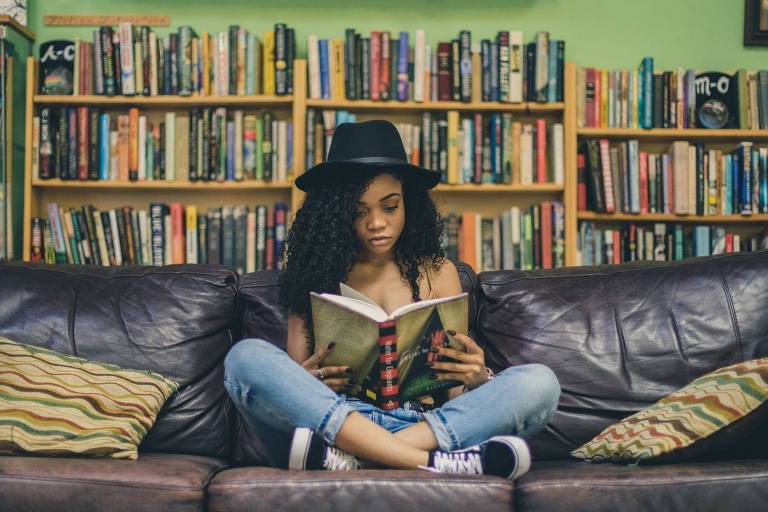 Garota lendo sentada no sofá usando chapéu com pernas cruzadas e biblioteca ao fundo
