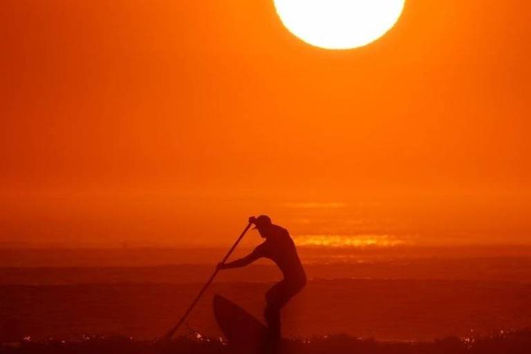 Imagem mostra um pôr do sol e um homem em cima de uma prancha segurando um remo