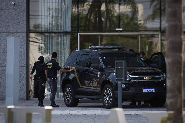 Dois policiais uniformizados estão ao lado de uma van preta da Polícia Federal, na entrada de um prédio
