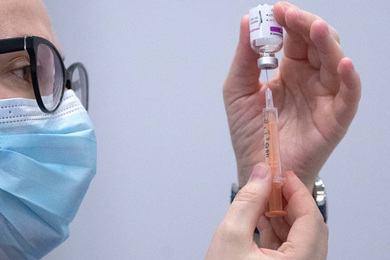 Imagem em close mostra uma pessoa de máscara e óculos espetando uma seringa em uma ampola de vacina