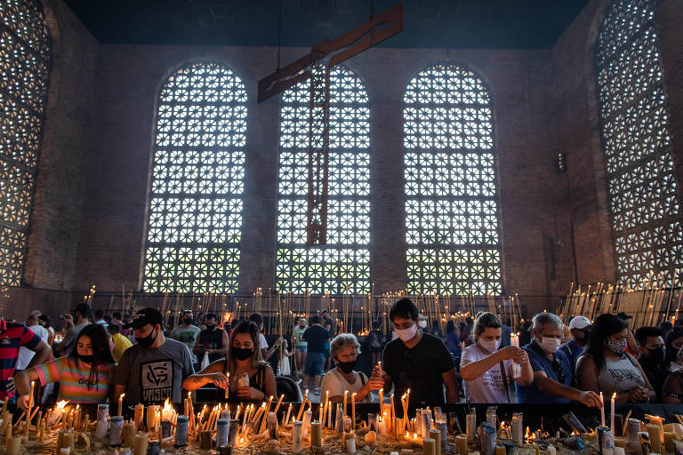 Imagem mostra bancada com velas acesas e fiéis em segundo plano, dentro de uma sala com pé direito alto, grandes janelas ao fundo e uma cruz pendurada no teto