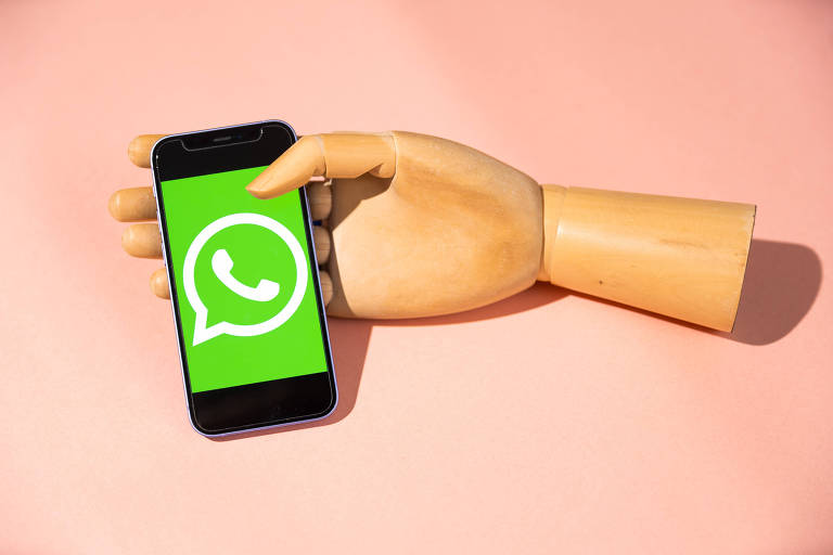 Na imagem, uma mão de manequim, de madeira, segura um smartphone preto. Na tela, está reproduzido o logotipo do WhatsApp, em verde e branco.