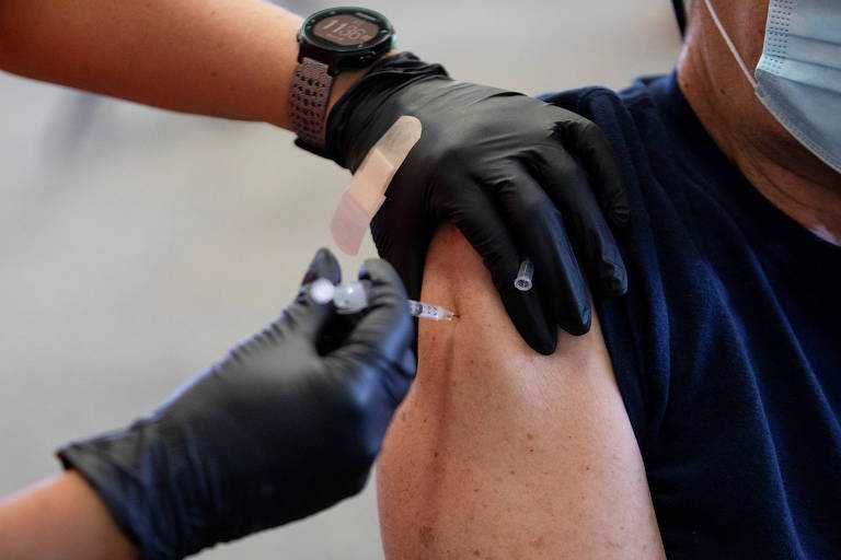 Mãos com luvas de borracha pretas aplicam injeção em um braço de pele branca, com camiseta azul marinho