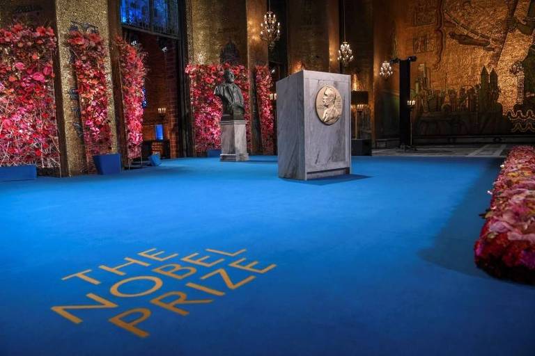 Em um chão azul se lê "the nobel prize". Ao fundo, há um púlpito.