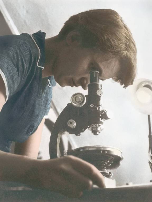 Uma mulher está com o corpo inclinando olhando para um microscópio