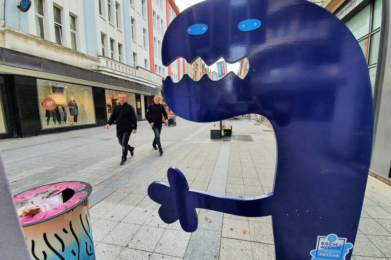 Pessoas caminham em rua comercial da cidade, vistas atrás de uma escultura de metal azul que representa um monstrinho