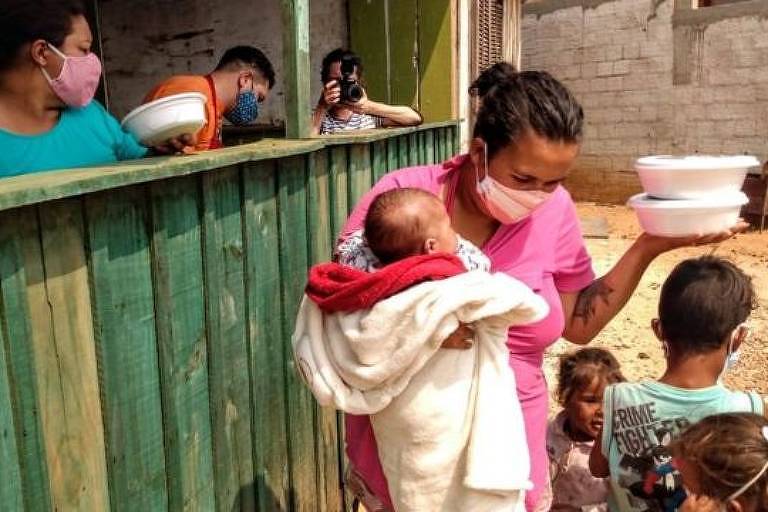 Duas mulheres seguram marmitas. Uma delas segura um bebê no colo e está próxima de três crianças