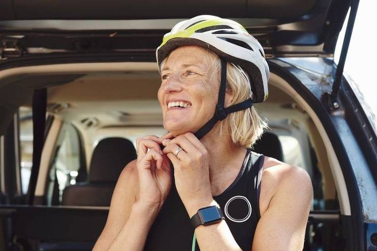 Imagem em primeiro plano mostra uma mulher usando capacete de ciclista. Ela sorri e leva as mãos para a altura do queixo. Ao fundo, se vê o porta malas de um carro aberto.