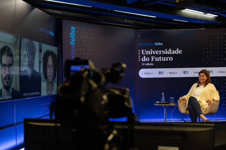 Repórter da folha, Erica Fraga, aparece sentada em uma cadeira no Auditório Folha. Ao lado, uma tela mostra a participação online de três convidados do debate