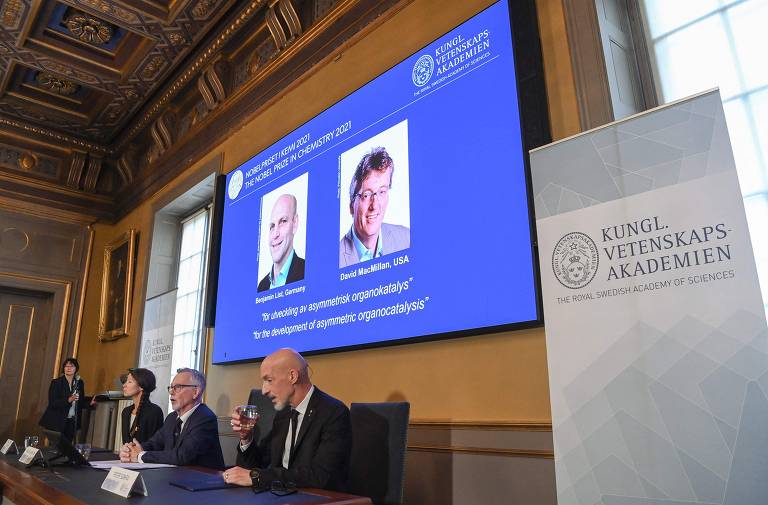 Em destaque em um telão, há a fotografia dos Cientistas Benjamin List e David MacMillan, vencedores do Nobel de Química. No canto inferior da imagem, há três pessoas sentadas de frente para uma mesa.