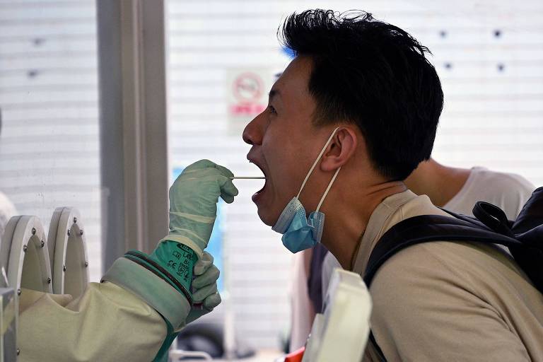 Um home está de perfil com a boca aberta e máscara no queixo. Na frente dele, um profissional da saúde coloca uma haste flexivel dentro da boca dele.