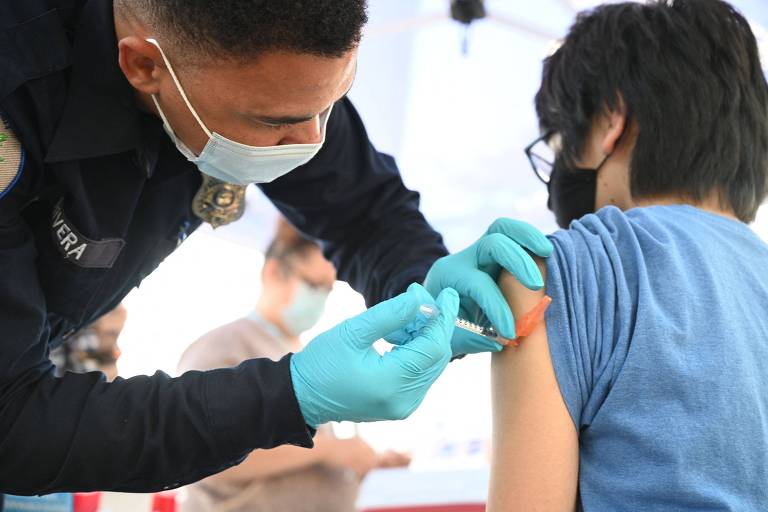 Profissional de saúde com casaco preto, máscara azul e luvas também azuis injeta vacina no braço de um jovem de camiseta azul e máscara preta