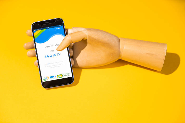 Uma mão de madeira está segurando um celular que exibe a tela inicial do aplicativo 'Meu INSS'. A tela do aplicativo mostra a mensagem 'Bem-vindo ao Meu INSS!' e os logotipos do INSS e do Governo Federal do Brasil. O fundo da imagem é amarelo.